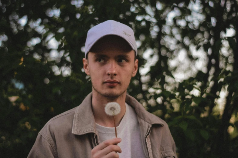 a man holding a dandelion in front of his face, an album cover, by Attila Meszlenyi, pexels contest winner, wearing a baseball cap, smooth pale skin, discord profile picture, portrait soft low light