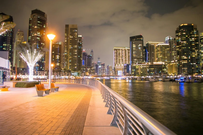 a bench sitting on top of a sidewalk next to a body of water, by Bernardino Mei, pexels contest winner, dubai, city led lights, high rise buildings, youtube thumbnail