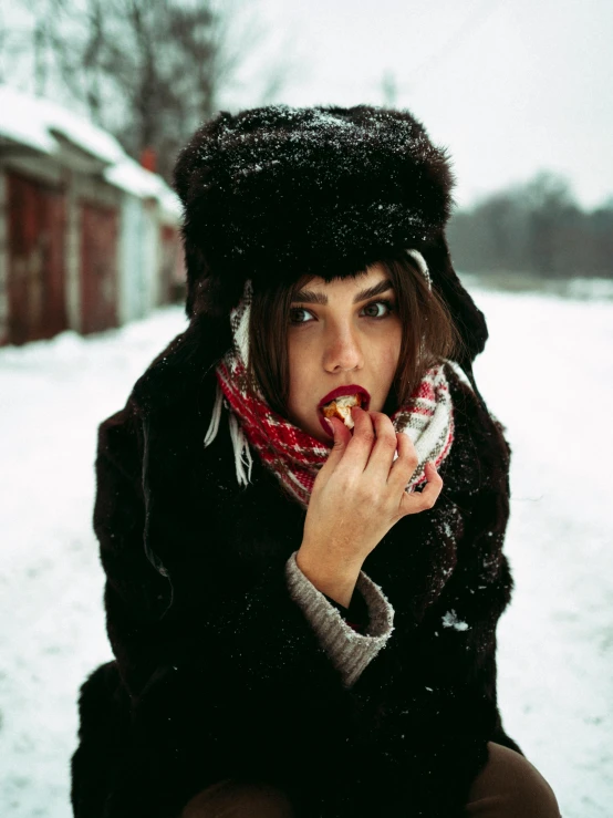 a woman sitting on a bench in the snow, an album cover, inspired by Elsa Bleda, pexels contest winner, tongue out, russian style, black hat, eating