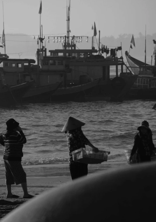 a group of people standing on top of a beach next to a body of water, a black and white photo, pexels contest winner, realism, fish market stalls, ships, :: morning, vietnamese woman