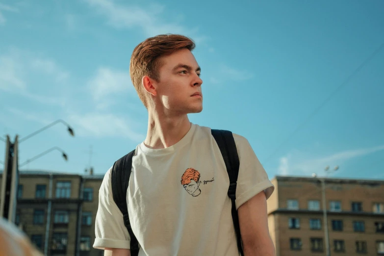 a man with a backpack standing in front of a building, a character portrait, by Julia Pishtar, trending on pexels, embroidered shirt, ginger hair, cute young man, dressed in a white t shirt