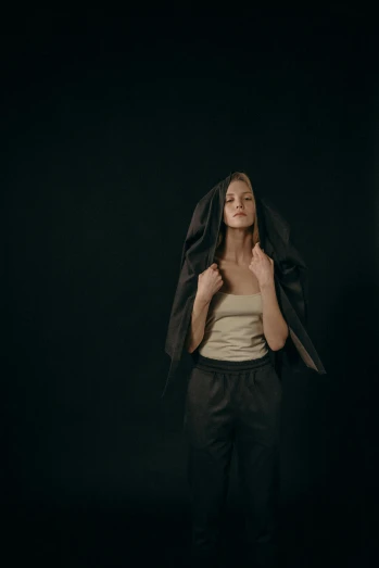 a woman standing in front of a black background, an album cover, inspired by Anna Füssli, pexels contest winner, wearing a hoodie and sweatpants, wearing a brown cape, frown fashion model, in an empty black room