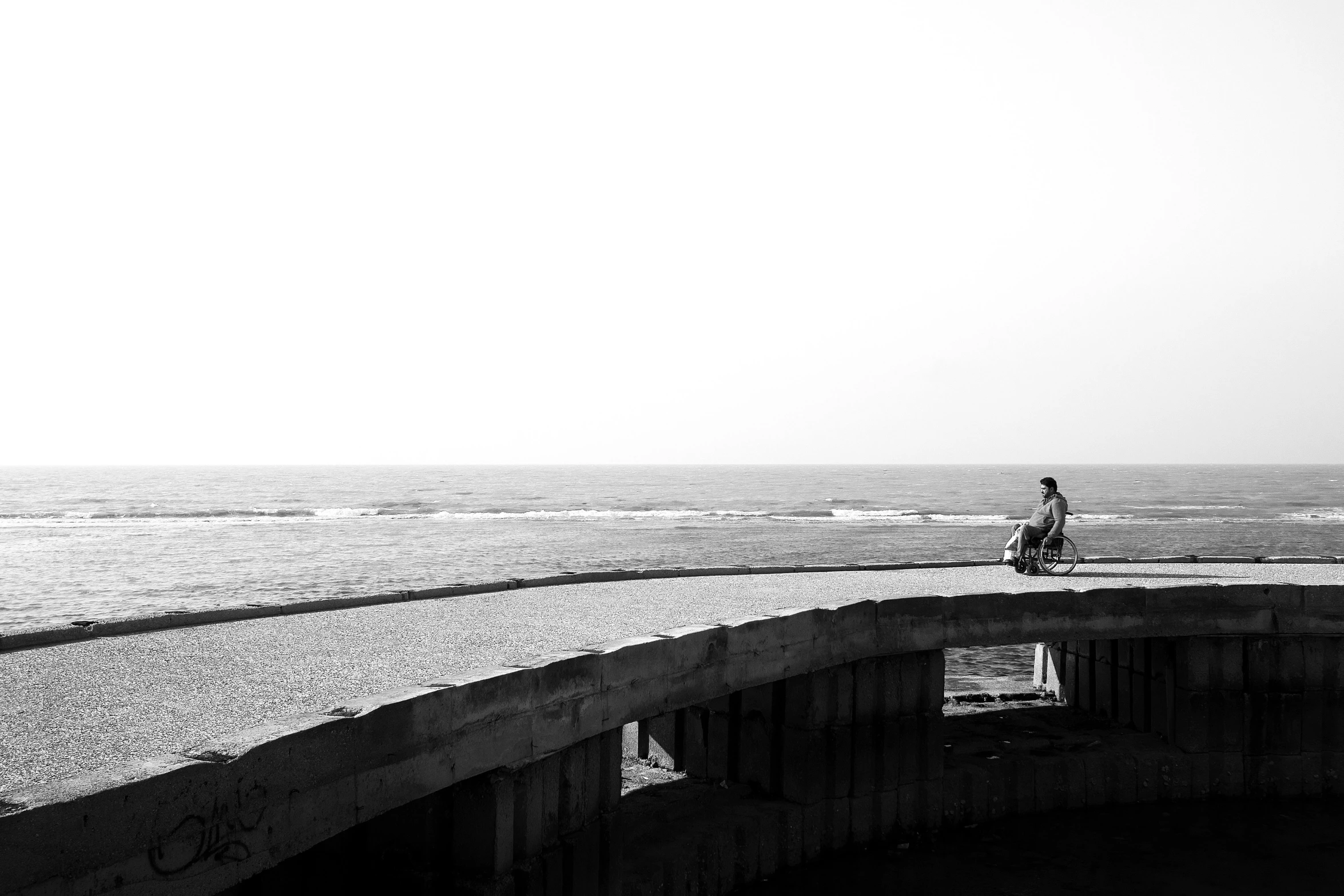 a man riding a bike across a bridge next to the ocean, a black and white photo, by Youssef Howayek, sitting alone, in sunny weather, sittin, nuri iyem