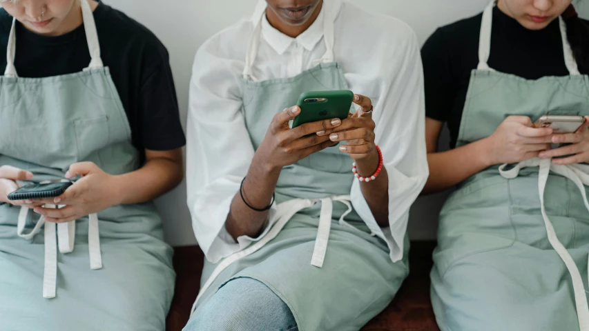 three people sitting on a bench looking at their cell phones, by Emma Andijewska, trending on pexels, happening, wearing an apron, in the kitchen, teal uniform, photo of a black woman