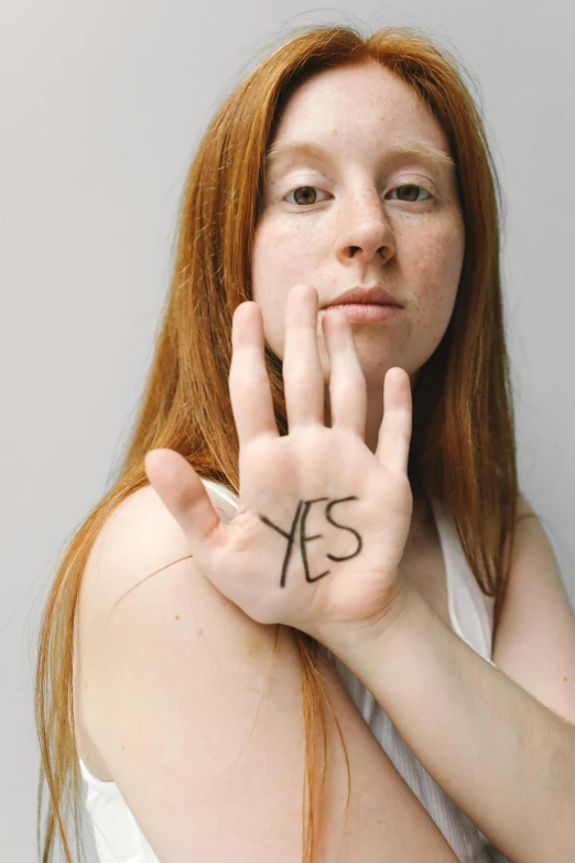 a woman holding up her hand with the word yes written on it, an album cover, inspired by Vanessa Beecroft, unsplash, renaissance, long ginger hair, patricia piccinini, prompt young woman, college