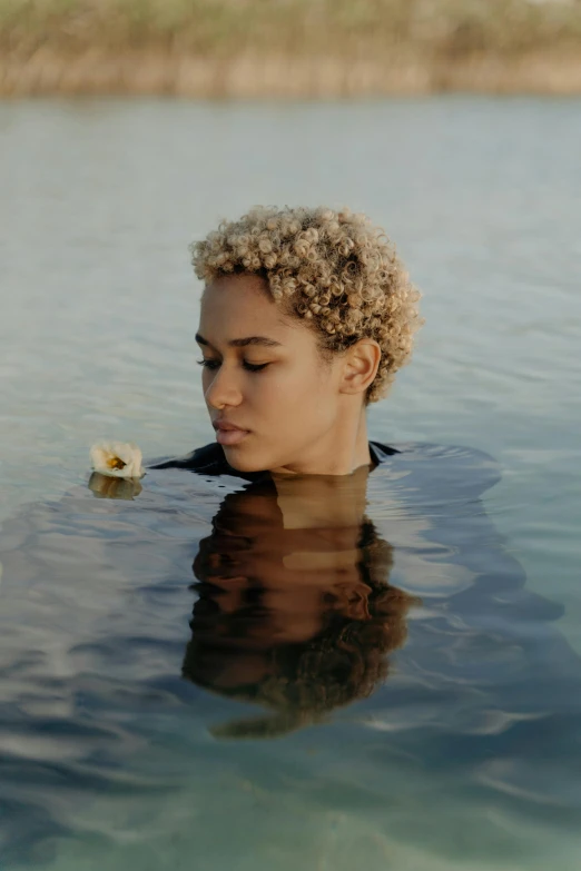 a woman in a body of water with a flower in her hand, short blonde afro, deep in thought, looking towards camera, brown skin like soil