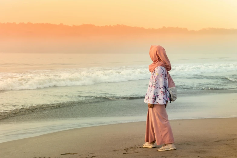 a woman standing on top of a beach next to the ocean, by Matija Jama, pexels contest winner, sumatraism, hijab, pink and orange, faded glow, casually dressed