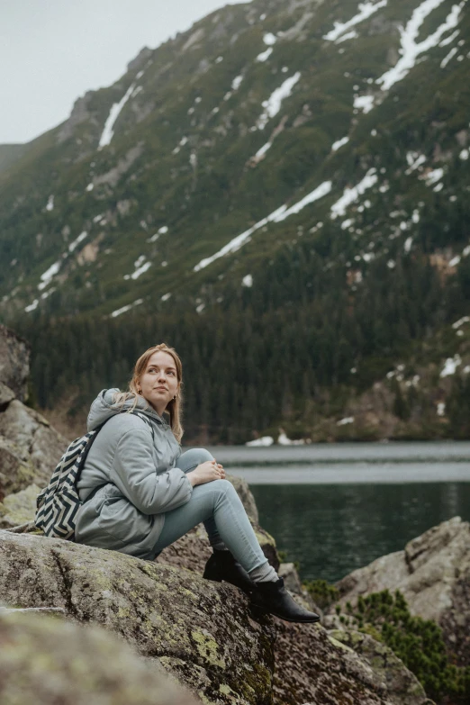 a woman sitting on top of a rock next to a lake, gray hoodie, with a backpack, jakub rebelka, profile image