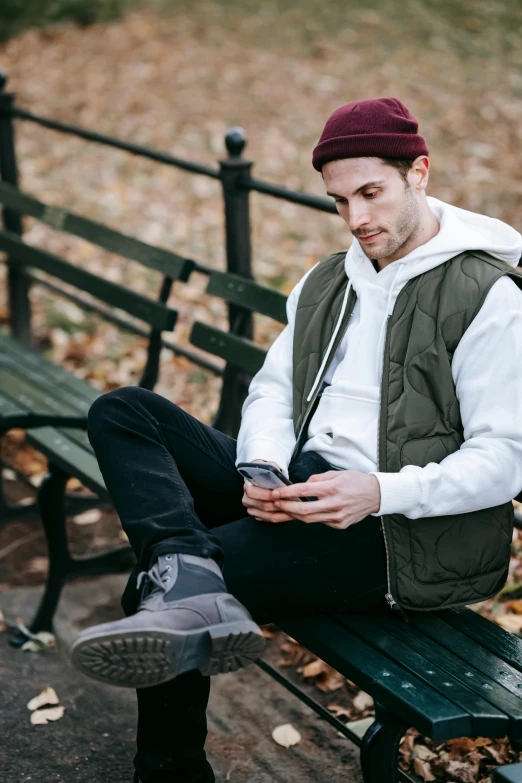 a man sitting on a bench looking at his cell phone, model wears a puffer jacket, wearing a vest, wearing a white sweater, owen gent