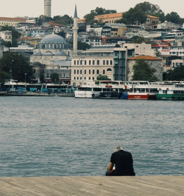a person sitting on a dock next to a body of water, by Yasar Vurdem, hurufiyya, entire city in view, mosque, cinematic footage, gray men