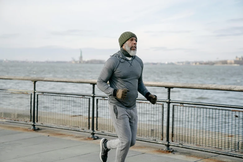 a man running on a sidewalk next to a body of water, he also wears a grey beanie, white beard, 2 0 2 2 photo, in new york city