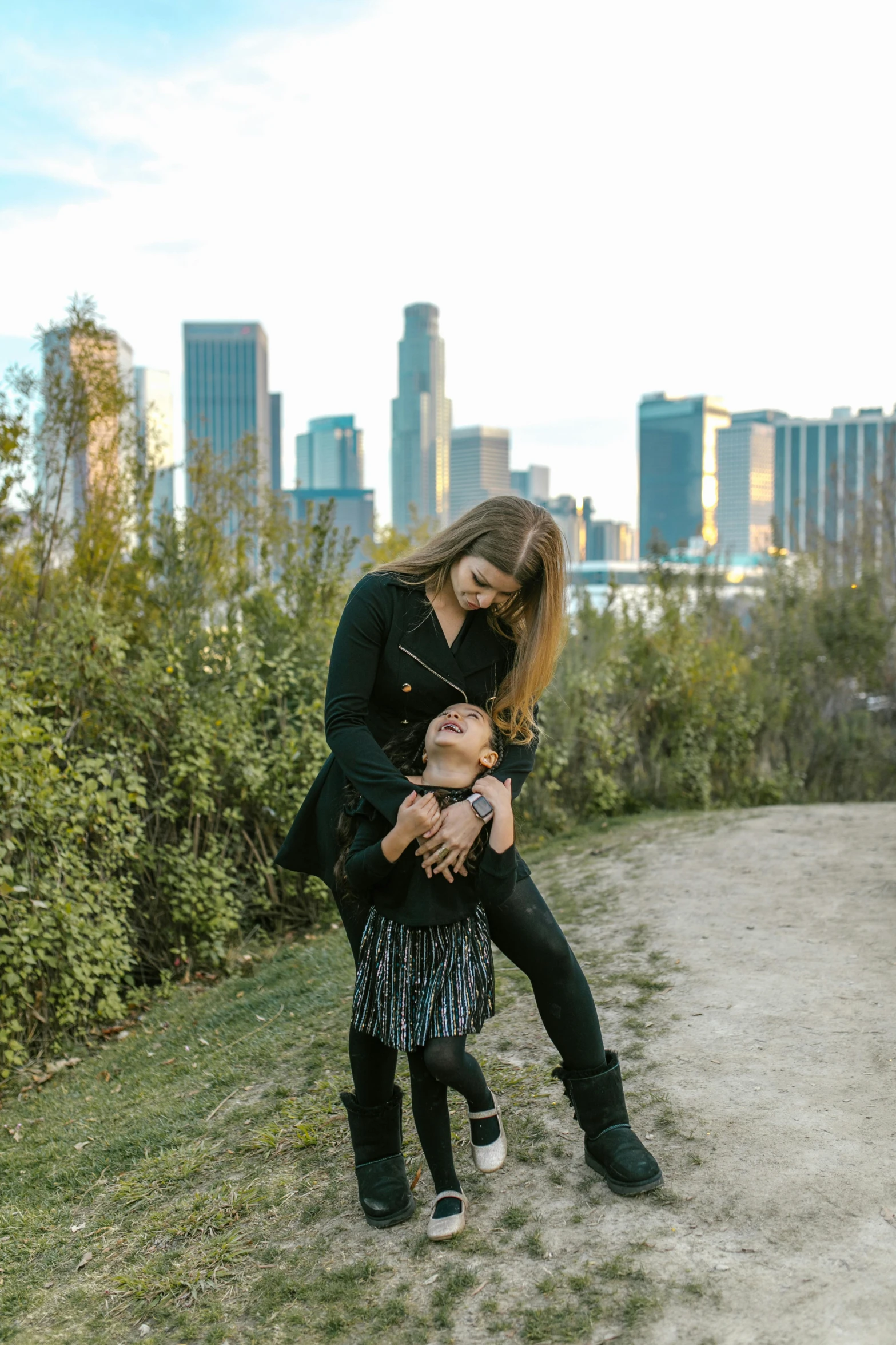 a woman holding a child on top of a dirt road, skyscrapers in the background, at a park, dasha taran, los angelos