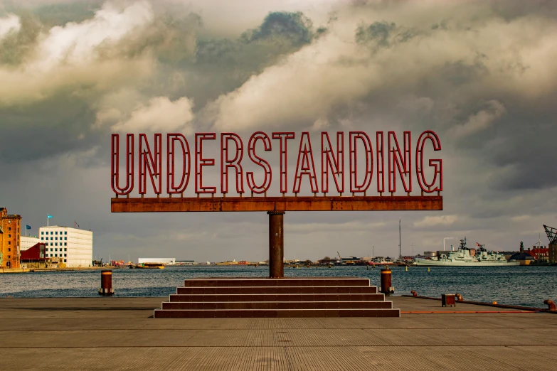 a large sign sitting in front of a body of water, by Jesper Knudsen, unsplash, hyperrealism, underground art, in a beachfront environment, knowledge, koyaanisqatsi