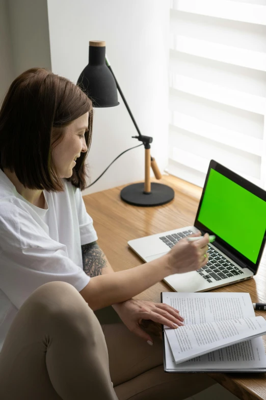 a woman sitting at a desk using a laptop with a green screen, studying in a brightly lit room, green and white, taken in the early 2020s, no - text no - logo