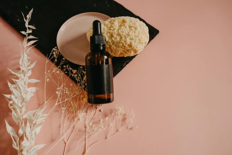 a bottle of essential oil next to a plate of flowers, trending on pexels, pink cloudy background, with textured hair and skin, cake, black oil