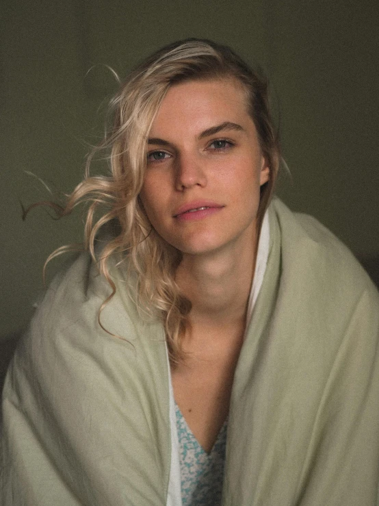a woman wrapped in a blanket sitting on a bed, inspired by Elsa Bleda, unsplash, photorealism, blond hair green eyes, julian ope, good looking face, low quality photo