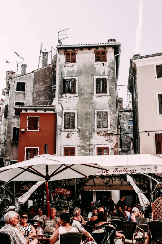 a group of people sitting at tables under umbrellas, a picture, trending on unsplash, romanticism, whitewashed buildings, burnt sienna and venetian red, crumbling buildings, seen from outside