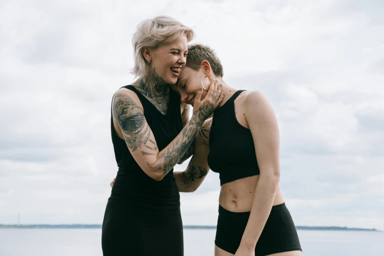 a couple of women standing next to each other on a beach, a tattoo, trending on pexels, antipodeans, laughing maniacally, a pale skin, skintight black clothes, portrait of women embracing