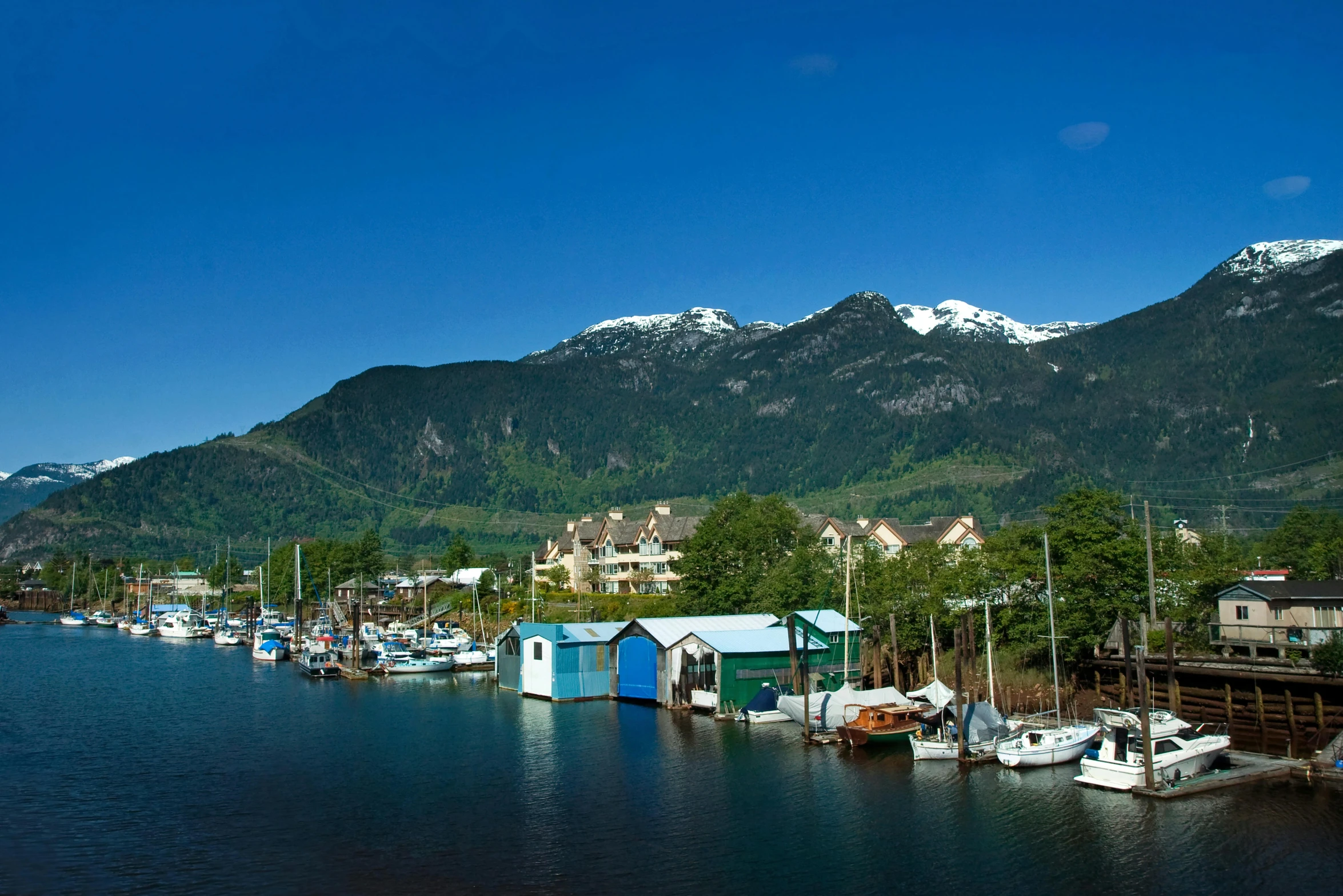 a number of boats in a body of water with mountains in the background, a picture, vancouver school, avatar image, waterfront houses, clear blue skies, conde nast traveler photo