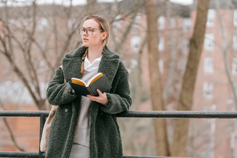 a woman standing in front of a fence holding a book, a portrait, inspired by Louisa Matthíasdóttir, trending on unsplash, realism, wearing a long coat, wearing reading glasses, college students, looking serious