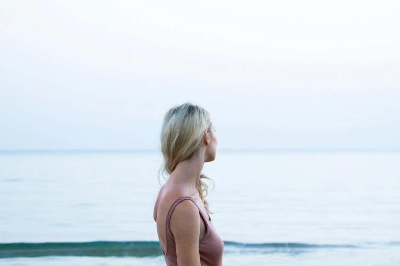 a woman standing on top of a beach next to the ocean, by Emma Andijewska, unsplash, wearing a camisole, side profile view, pink, slight overcast lighting