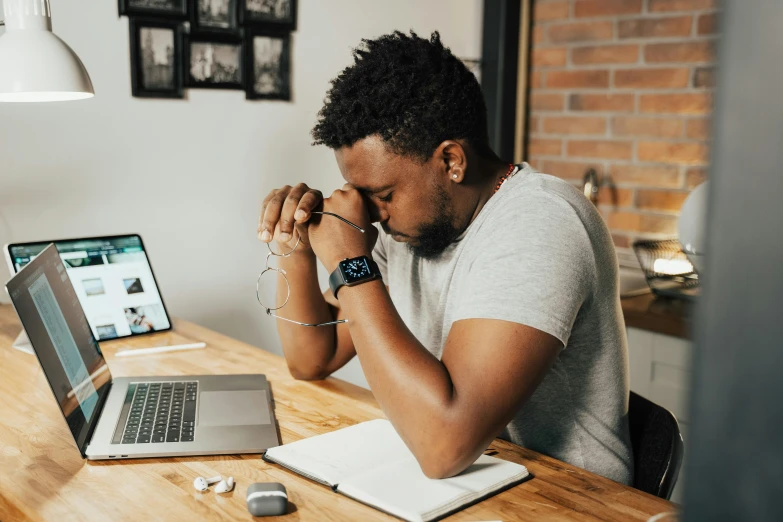 a man sitting at a table in front of a laptop, by Carey Morris, trending on pexels, renaissance, praying, looking exhausted, black man, in an office