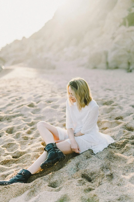 a woman sitting on top of a sandy beach, inspired by Elsa Bleda, unsplash contest winner, white blouse and gothic boots, pale glowing skin, zoe kazan, white tunic