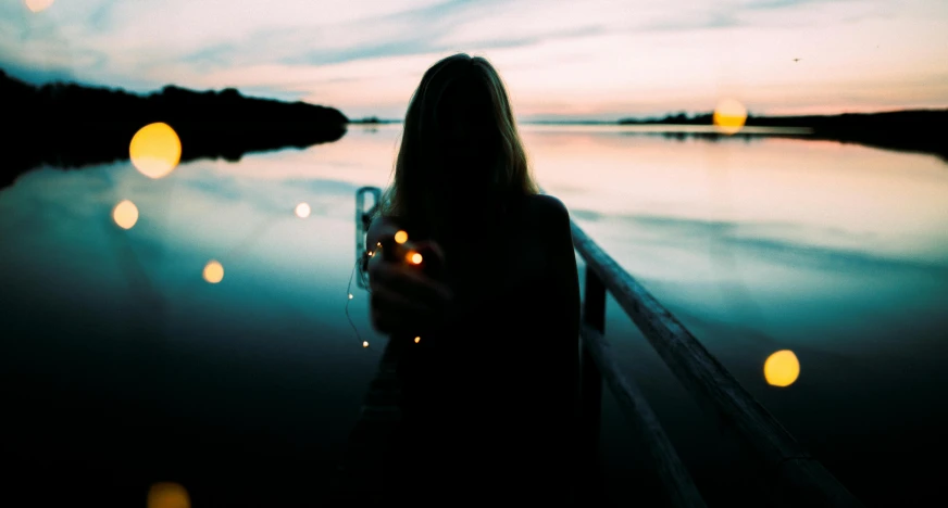 a woman standing on a dock holding a glass of wine, inspired by Elsa Bleda, pexels contest winner, floating lights, eerie person silhouette, instagram post, unfocused