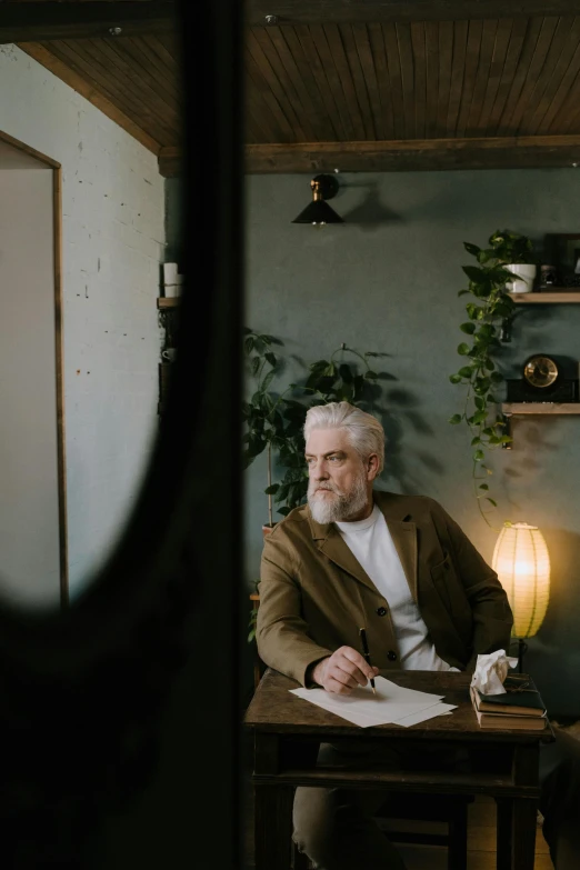 a man sitting at a table in a room, a character portrait, pexels contest winner, gray beard, wood and paper, distant photo, well lit professional photo
