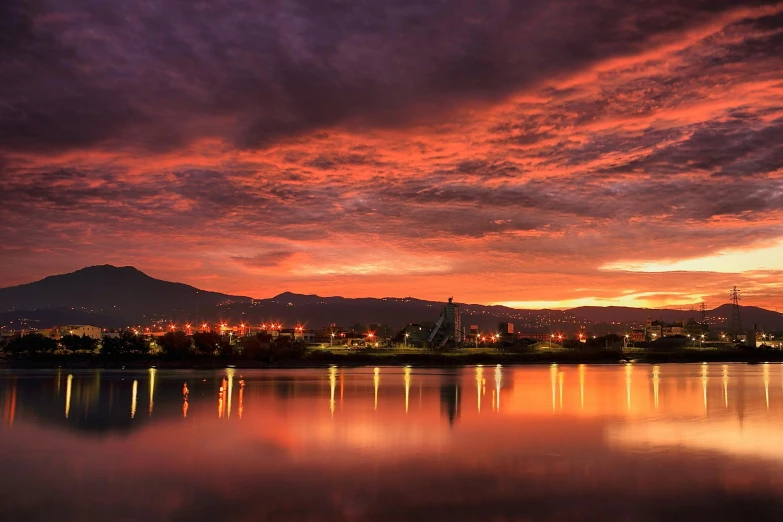 a sunset over a body of water with mountains in the background, city light reflections, orange and red lighting, industrial photography, new zeeland