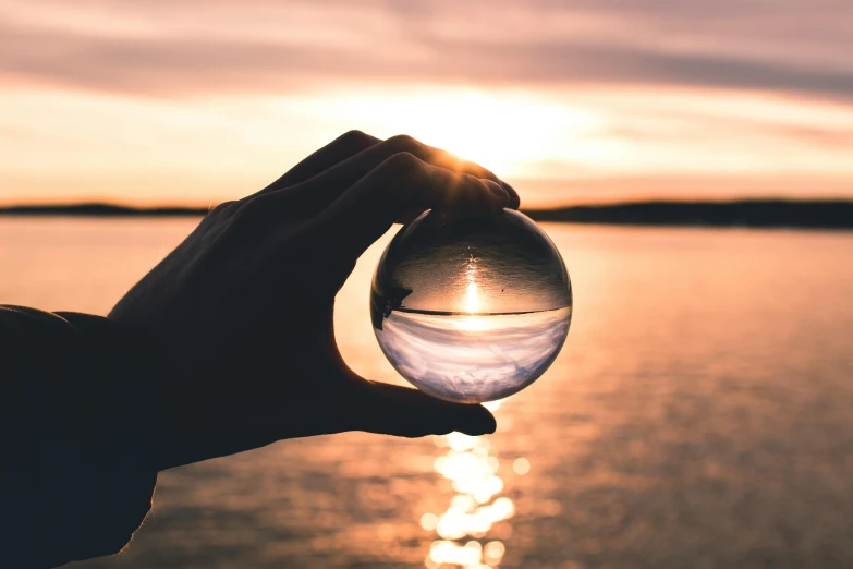 a person holding a glass ball in front of a body of water, with a sunset