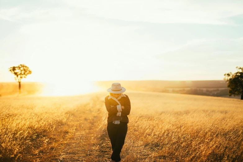 a person standing in a field with a hat on, unsplash, golden glow, walking down, traveller, trailing off into the horizon