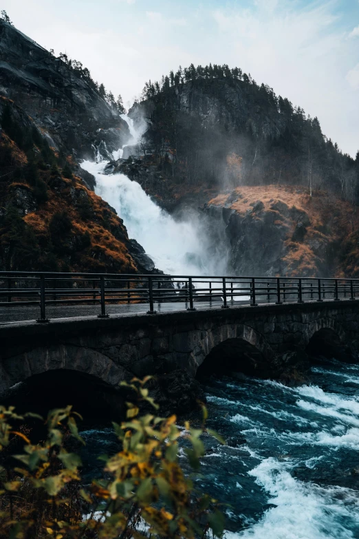 a bridge over a river with a waterfall in the background, by Johannes Voss, pexels contest winner, turbulent lake, mid fall, mountainside, thumbnail