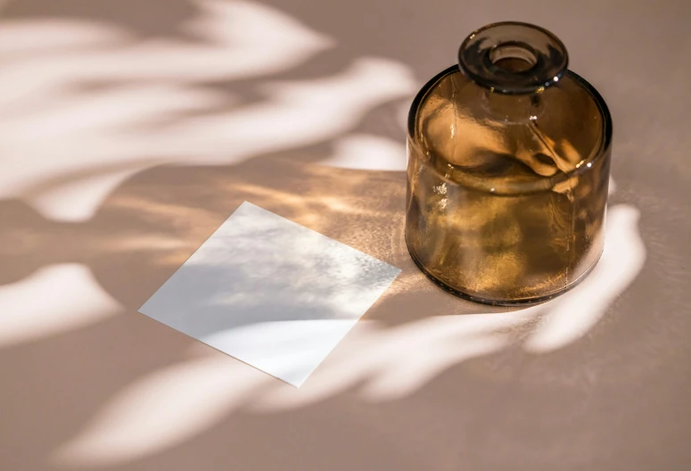 a glass jar sitting on top of a table next to a piece of paper, an album cover, inspired by Agnes Martin, unsplash, light and space, sun and shadow, square sticker, shadow play, gradient brown to white