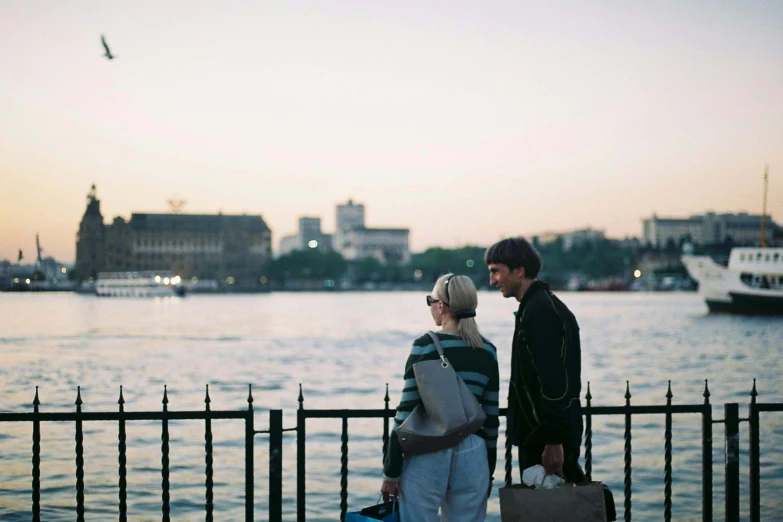 a couple standing next to each other near a body of water, happening, river thames, early evening, traveller, easygoing