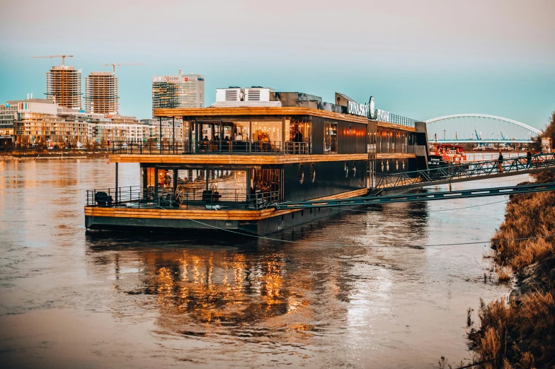 a boat sitting on top of a river next to a bridge, by Julia Pishtar, pexels contest winner, bauhaus, restaurant exterior photography, gold glow, memphis, three - quarter view