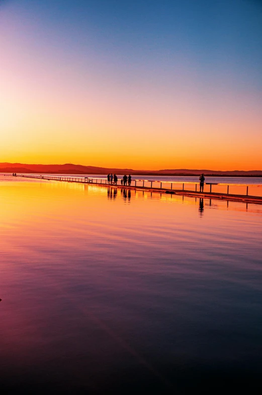 a group of people standing on top of a beach next to a body of water, a picture, sunrise colors, manly, boardwalk, smooth reflections