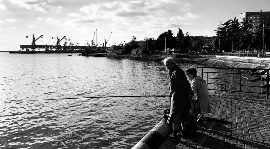 a couple of people standing next to a body of water, a black and white photo, by Mathias Kollros, harbor, fishing, parents watching, 🪔 🎨;🌞🌄