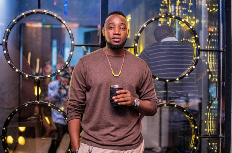 a man standing in front of a window holding a cup, an album cover, by Chinwe Chukwuogo-Roy, pexels contest winner, in a nightclub, smol, confident looking, mkbhd