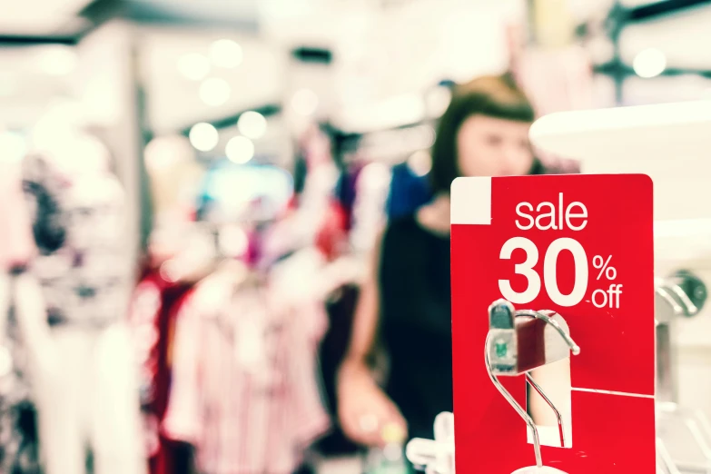 a sale sign in a store with a woman in the background, a photo, by Julia Pishtar, shutterstock, brand colours are red and blue, loosely cropped, people shopping, 🎀 🗡 🍓 🧚