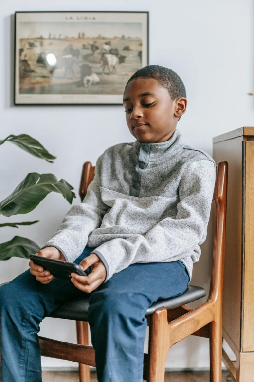 a boy sitting in a chair holding a tablet, by Everett Warner, trending on pexels, wearing a sweater, black teenage boy, holding a very advance phone, in a living room