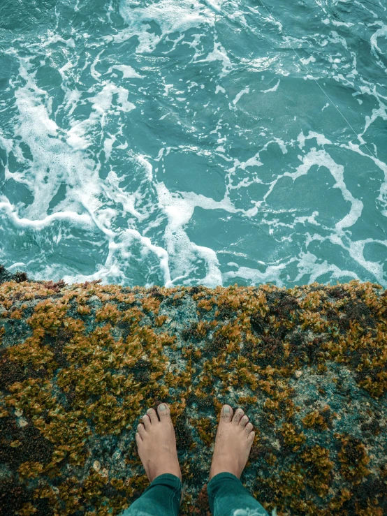 a person standing on top of a rock next to the ocean, inspired by Elsa Bleda, pexels contest winner, happening, algae feet, high view, teal aesthetic, wall of water either side