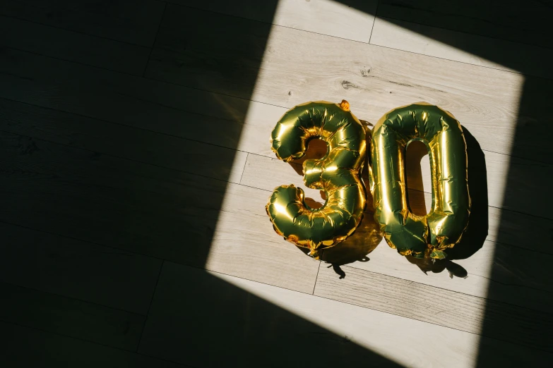 a number 30 balloon sitting on top of a wooden floor, by Emma Andijewska, trending on unsplash, gold foil, birthday cake on the ground, an elderly, paul barson