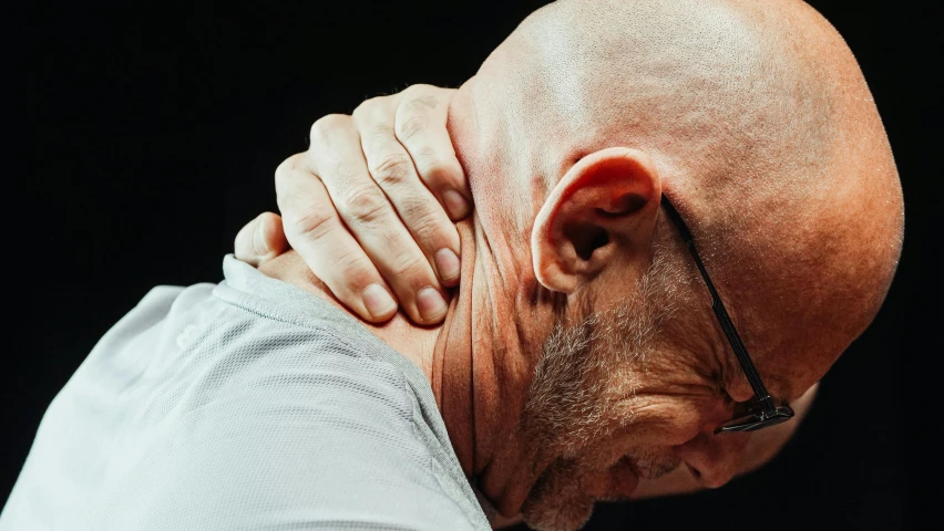 a bald man with glasses rubbing his neck, a photo, unsplash, in pain, side view close up of a gaunt, profile image, his back is turned