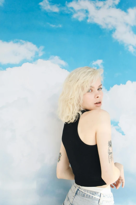 a woman standing against a wall with clouds in the background, an album cover, featured on reddit, pale hair, cumulus tattoos, pale fair skin!!, catalog photo