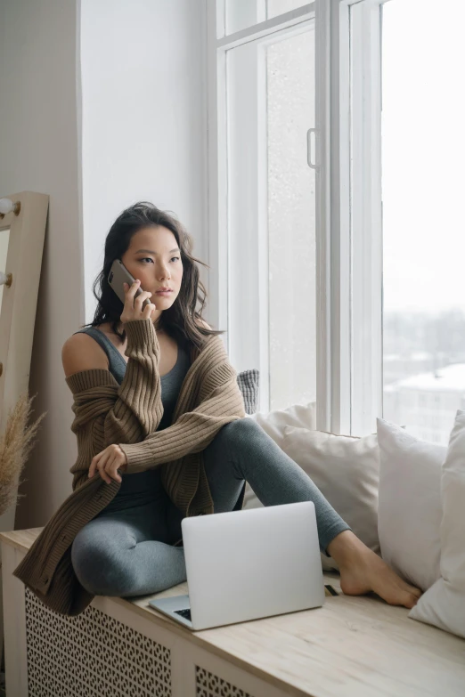 a woman sitting on a window sill talking on a cell phone, inspired by helen huang, trending on pexels, minimalism, in front of a computer, brown clothes, joanna gaines, asian human