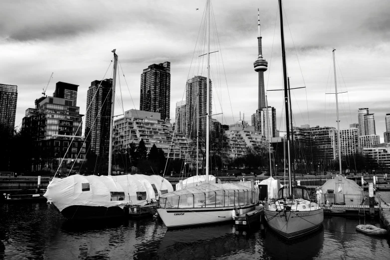 a number of boats in a body of water with a city in the background, a black and white photo, by Seb McKinnon, pexels contest winner, cn tower, waterfront houses, wooden sailboats, white pale concrete city