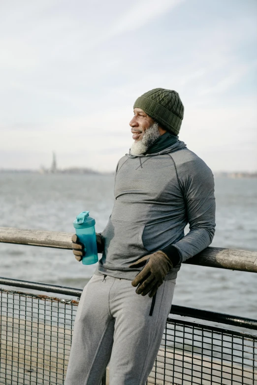 a man standing next to a body of water, holding a bottle, cold weather, wearing fitness gear, gray beard