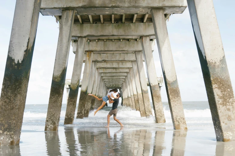 a man carrying a woman under a pier, by Josh Bayer, unsplash contest winner, visual art, playful pose of a dancer, concrete pillars, in a beachfront environment, an intricate