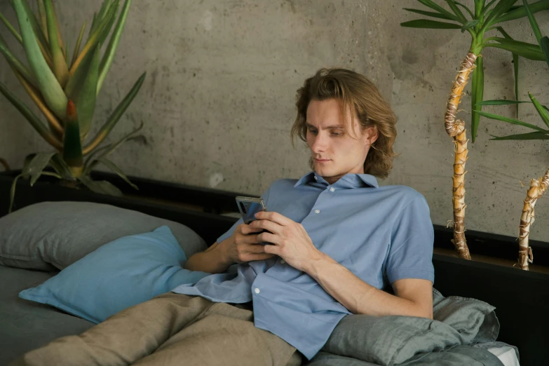 a man sitting on a bed looking at his cell phone, by Alice Mason, trending on pexels, happening, wearing a light blue shirt, sitting in a lounge, nikolay georgiev, avatar image